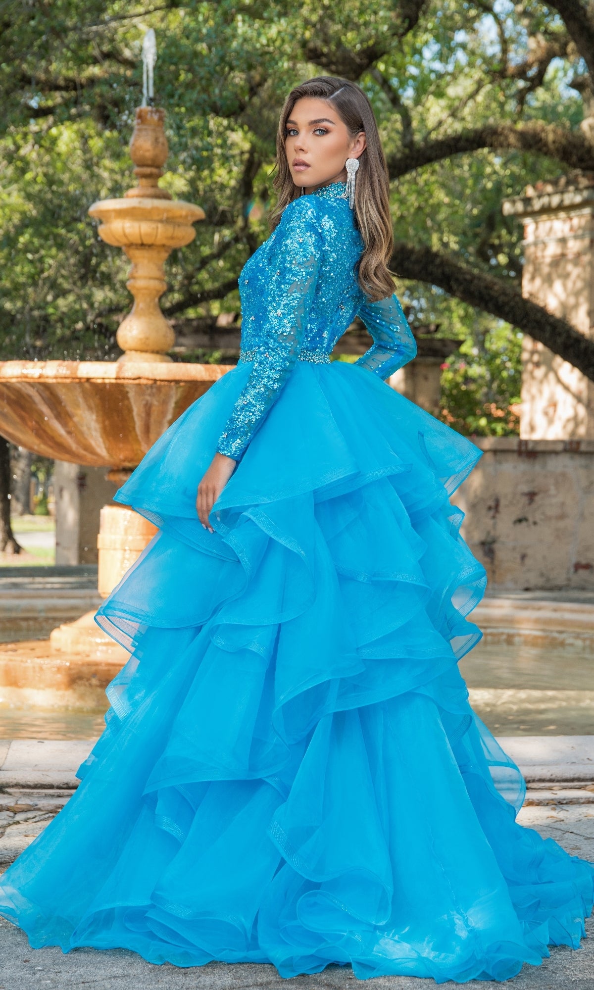 Woman wearing the Ava Presley 28595 Ruffle Short Sequin Romper in bright blue, posing outdoors by a stone fountain and trees.